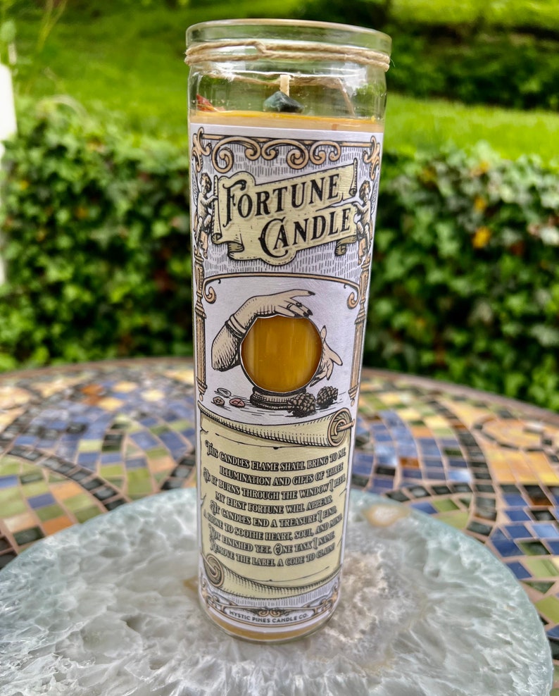 May include: A fortune candle with a yellow wax and a crystal embedded in the top. The candle is in a glass jar with a label that says "Fortune Candle" and has a quote about the candle's flame bringing hope, illumination, and gifts of their dreams. The label also says "Mystic Pines Candle Co."