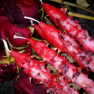 May include: Close-up of four red taper candles with white wicks, surrounded by dried red rose petals and stems. The candles have a textured appearance with visible wax drips and embedded floral elements.