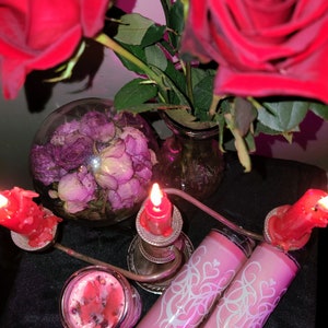 May include: Two pink candles with white designs and a heart motif, two red candles in silver candle holders, and a glass bowl filled with dried pink roses.