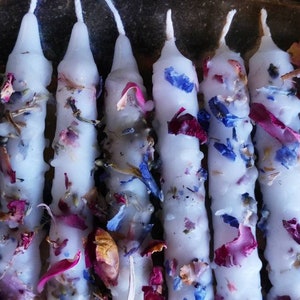 May include: A close-up of a group of white candles with dried flowers and petals embedded in the wax. The flowers are pink, blue, and brown.