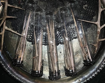 May include: A collection of clear glass vials, each containing a long, sharp, brown thorn. The vials are arranged on a decorative silver tray, with additional thorns placed around the edges. The image has a dark, gothic aesthetic.