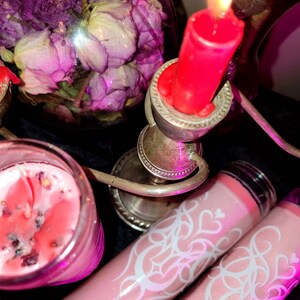 May include: Three pink candles with white designs. One candle is in a glass jar with a red wax top. The other two candles are in glass tubes with a pink wax top. The candles are on a silver candlestick with a red candle on top.