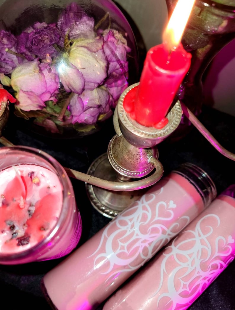 May include: Three pink candles with white designs. One candle is in a glass jar with a red wax top. The other two candles are in glass tubes with a pink wax top. The candles are on a silver candlestick with a red candle on top.