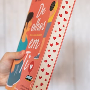 以下が含まれることがあります： 「De olhos em Ti」というタイトルとハートのシンボルがついたピンク色のペーパーバック。本の表紙には、女性と男性のイラストが描かれている。本の背表紙は白地に赤いハートが描かれている。