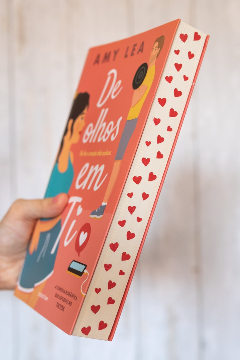 以下が含まれることがあります： 「De olhos em Ti」というタイトルとハートのシンボルがついたピンク色のペーパーバック。本の表紙には、女性と男性のイラストが描かれている。本の背表紙は白地に赤いハートが描かれている。