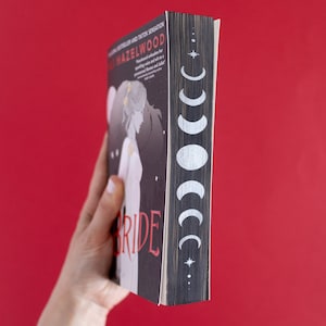 Puede incluir: Un libro de bolsillo con una cubierta negra y texto blanco que dice "The Bride" con un dise&ntilde;o de luna y estrella en el lomo. El libro est&aacute; sostenido en una mano.