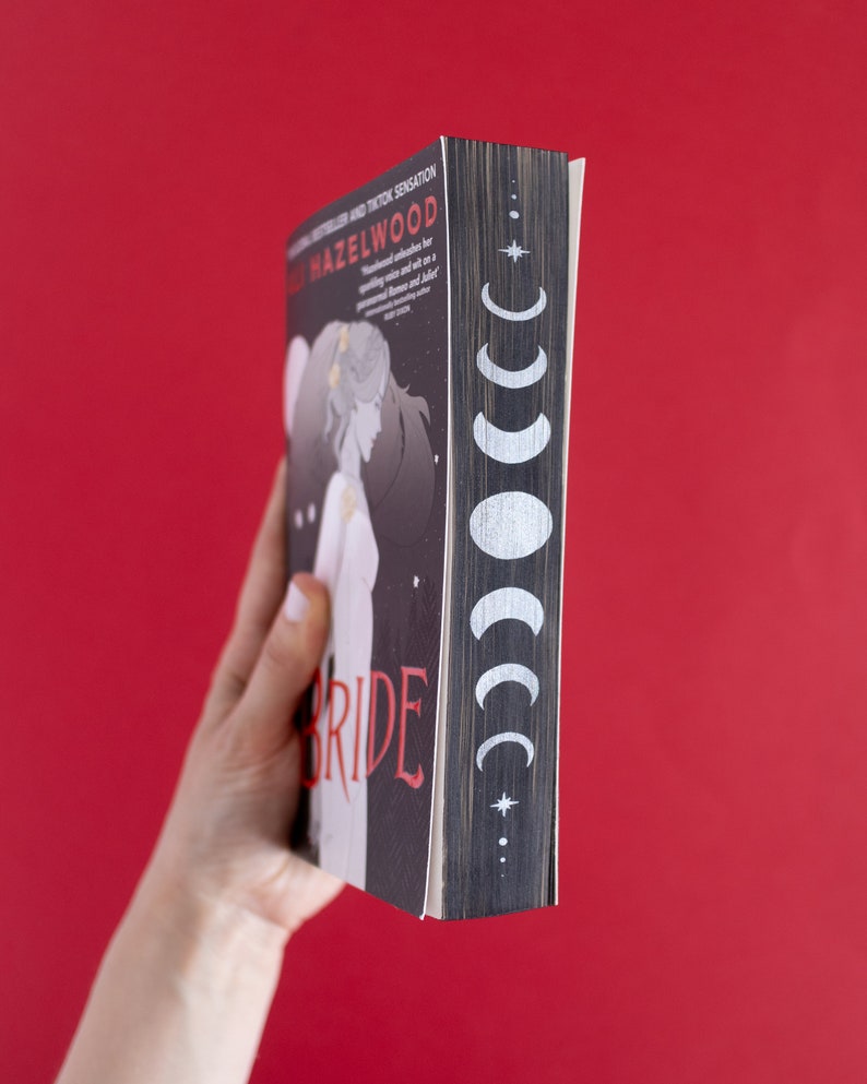 Puede incluir: Un libro de bolsillo con una cubierta negra y texto blanco que dice "The Bride" con un dise&ntilde;o de luna y estrella en el lomo. El libro est&aacute; sostenido en una mano.