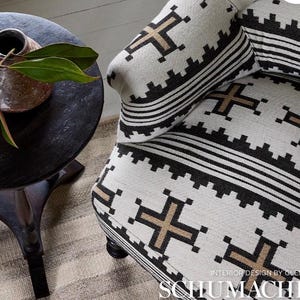 May include: Close-up of a patterned armchair with a matching pillow. The fabric features a repeating geometric design in black and tan against a cream background. A dark wooden side table with a vase and greenery is in the background.