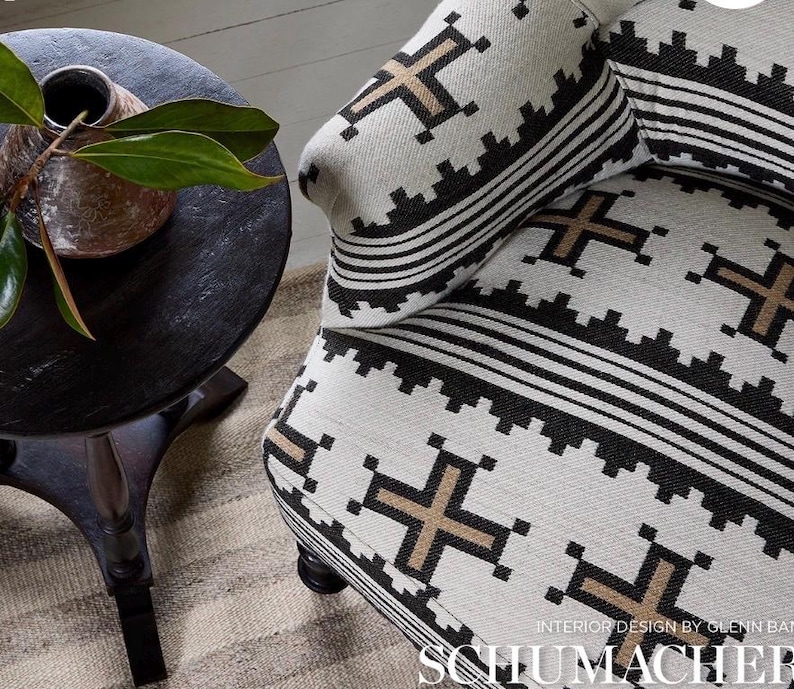 May include: Close-up of a patterned armchair with a matching pillow. The fabric features a repeating geometric design in black and tan against a cream background. A dark wooden side table with a vase and greenery is in the background.
