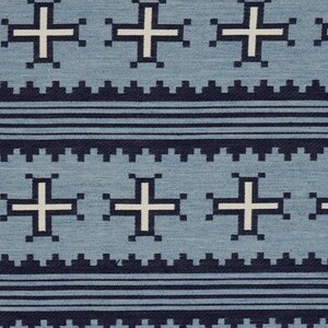 May include: Close-up of a blue fabric with a repeating pattern. The design features white crosses and dark blue geometric shapes. Horizontal dark blue lines and a zigzag border complete the design.