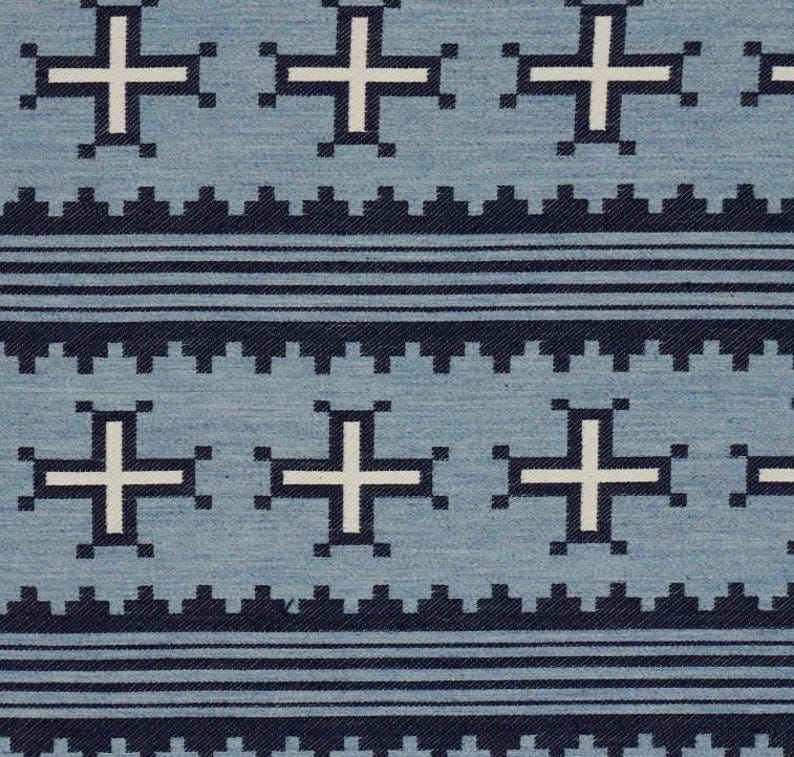 May include: Close-up of a blue fabric with a repeating pattern. The design features white crosses and dark blue geometric shapes. Horizontal dark blue lines and a zigzag border complete the design.