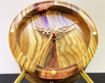 May include: A wooden clock with a copper-colored face and hands. The clock is made of a light brown wood with dark brown streaks. The clock has a round face and three legs. The clock is sitting on a black surface.