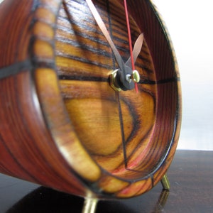 Puede incluir: Un reloj de madera con un acabado de madera de grano rojo y marrón. El reloj tiene una esfera negra con manecillas doradas y una perilla dorada. El reloj está en un soporte con tres patas doradas.