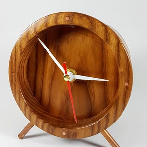 May include: A wooden clock with a round face and copper accents. The clock has a white face with black hour and minute hands and a red second hand. The clock is sitting on three copper legs.