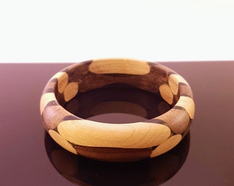 May include: A wooden bangle bracelet with a brown and light brown striped pattern. The bracelet is made of multiple pieces of wood that have been glued together to create a unique design.