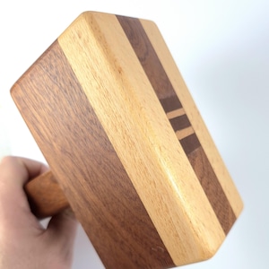 May include: A wooden mallet with a rectangular head and a round handle. The head is made of alternating dark and light wood stripes, with a small decorative stripe. The mallet is held by a hand against a white background.