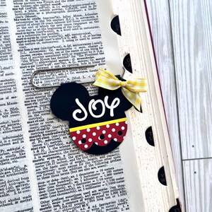 May include: A black and red Mickey Mouse-shaped ornament with white polka dots and the word "Joy" written in white letters. The ornament has a yellow and white gingham bow.