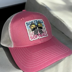 May include: Pink and white trucker hat with a softball and bow graphic. The graphic says "Softball" multiple times in different colours.