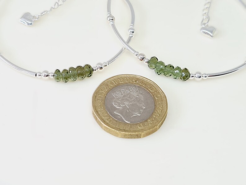 Peut inclure: Deux bracelets en argent avec des perles de pierres pr&eacute;cieuses vertes. Chaque bracelet a un design incurv&eacute; avec de petites perles argent&eacute;es et un charm en forme de c&oelig;ur. Une pi&egrave;ce d'or est au premier plan.
