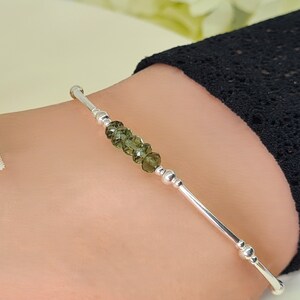 Peut inclure: Un bracelet en argent orn&eacute; d'une rang&eacute;e de perles de pierres pr&eacute;cieuses vert olive. Le bracelet a un design simple et &eacute;l&eacute;gant avec de petites perles d'argent et une finition lisse et polie. Le bracelet est pr&eacute;sent&eacute; sur un poignet.