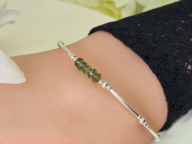 Peut inclure: Un bracelet en argent orn&eacute; d'une rang&eacute;e de perles de pierres pr&eacute;cieuses vert olive. Le bracelet a un design simple et &eacute;l&eacute;gant avec de petites perles d'argent et une finition lisse et polie. Le bracelet est pr&eacute;sent&eacute; sur un poignet.