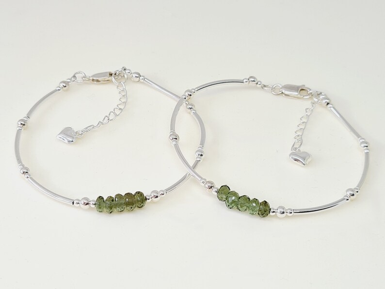 Peut inclure: Deux bracelets argent&eacute;s d&eacute;licats. Chaque bracelet pr&eacute;sente une rang&eacute;e de petites pierres pr&eacute;cieuses vert olive et des perles argent&eacute;es. Les bracelets ont des fermoirs r&eacute;glables et un petit charme en forme de c&oelig;ur.