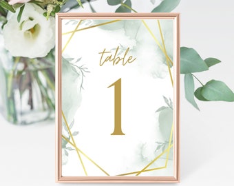 May include: A white table number card with a gold geometric frame and green watercolor accents. The card says "table 1" in gold lettering.