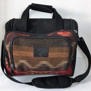 May include: Black travel bag with a brown, red and beige patterned fabric exterior. The bag has a top handle and a shoulder strap. The fabric has a southwestern design.
