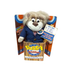 Puede incluir: Un juguete de peluche con forma de león, con una chaqueta azul y un botón de campaña. El juguete está en una caja de madera con la inscripción "Bubba for President" y un letrero que dice "Try Me!" y "Hold my mouth to try and keep me quiet!"