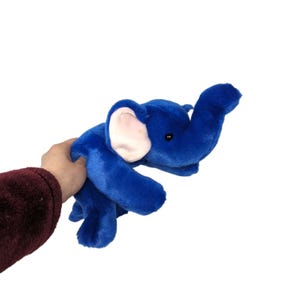 Op de afbeelding: Een blauw pluche olifantenspeelgoed met lichtroze binnenoren en kleine zwarte ogen. De olifant is gemaakt van zacht, donzig materiaal en wordt tegen een witte achtergrond gehouden.