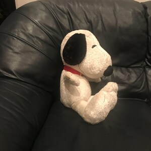 Peut inclure: Une peluche Snoopy blanche avec des oreilles et un nez noirs, portant un collier rouge. Le jouet en peluche est assis sur un canapé en cuir noir. Ce personnage de dessin animé classique est un jouet populaire.