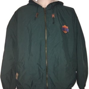 May include: A dark green hooded jacket with a full zipper closure. The jacket features a light-colored lining, a logo patch on the chest, and brown leather accents on the hood. The jacket has a drawstring at the hood and a tag on the lower left side.