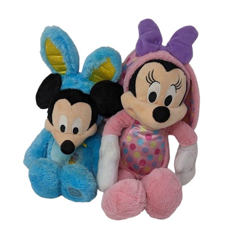 May include: Two plush Easter bunny Mickey Mouse characters. The Mickey Mouse on the left is wearing a blue bunny costume with yellow ears. The Mickey Mouse on the right is wearing a pink bunny costume with purple ears and a bow.