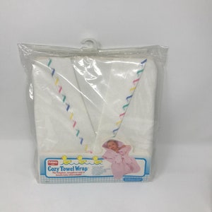 Könnte beinhalten: Ein weißes Baby-Handtuch in einer transparenten Plastikverpackung. Das Handtuch hat eine Kapuze und farbige Verzierungen. Die Verpackung zeigt den Text "Cozy Towel Wrap" und eine Abbildung eines Babys in einem rosa Handtuch.