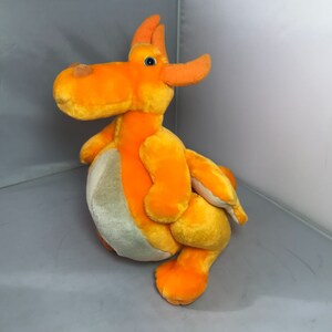 Pode incluir: Brinquedo de pelúcia de dragão laranja com um olho azul claro. O dragão tem uma barriga cinza claro e uma cauda longa.