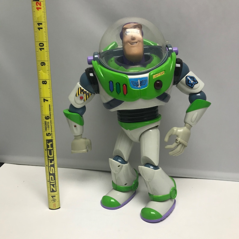 May include: A green and white Buzz Lightyear action figure with a clear plastic helmet and a red button on the chest. The figure has articulated arms and legs and is standing on a white surface.