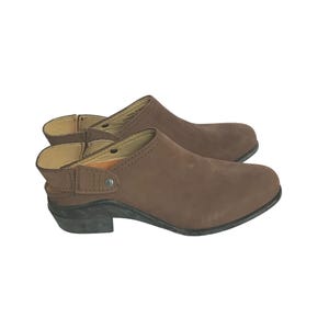 May include: A pair of brown suede clogs with a low heel. The clogs have a closed-toe design and an adjustable strap around the ankle. The soles are black, and the clogs appear to be unworn.
