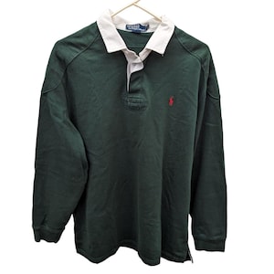 May include: Dark green long-sleeved rugby shirt with a white collar and a red logo on the chest. The shirt has a button-up placket and is hanging on a white hanger. The shirt is made of a soft, comfortable material.