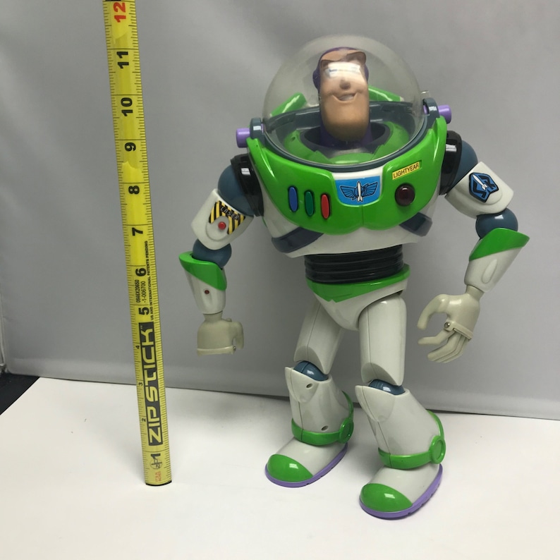 May include: A green and white Buzz Lightyear action figure with a clear plastic helmet and a red button on the chest. The figure has a purple and green base and is standing on a white surface.