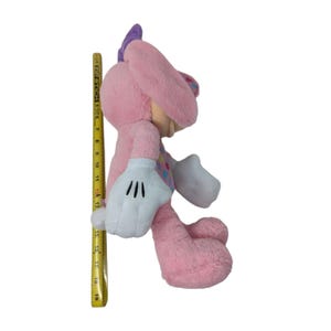 May include: Pink plush toy with a purple bow, white gloves, and colorful polka dots. The toy is sitting on a white background.