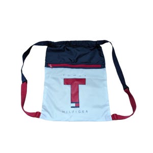 Puede incluir: Mochila con cordón azul marino y blanca con cremallera y correas rojas. La bolsa presenta un gran logotipo "T" rojo con la palabra "HILFIGER" impresa debajo. Diseñada para transportar artículos.