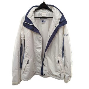 May include: A white and navy blue Columbia Sportswear jacket. The jacket has a hood, a zipper closure, and multiple pockets. The Columbia logo is visible on the sleeve. The jacket is hanging on a black hanger.