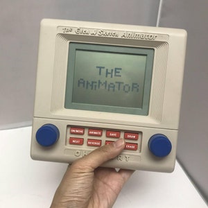 May include: A white Etch A Sketch Animator toy with blue knobs. The screen displays the text "THE ANIMATOR". The toy has buttons labelled "ON/MOVE", "ANIMATE", "SAVE", "NEXT", "REVERSE", "DRAW", and "ERASE".