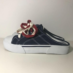 Peut inclure: Baskets slip-on en toile bleu marine avec semelles et embouts blancs. Les chaussures présentent des coutures blanches et des lacets rouges et blancs noués en nœud. L'arrière de la chaussure est ouvert.
