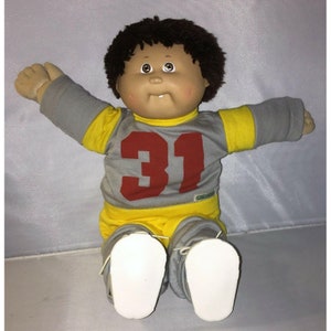 Può includere: Bambola Cabbage Patch Kids, con carnagione abbronzata, capelli castani ricci e un'espressione sorridente. La bambola indossa una maglia grigia e gialla con il numero 31 in rosso e pantaloni grigi abbinati. Le scarpe bianche completano l'outfit.