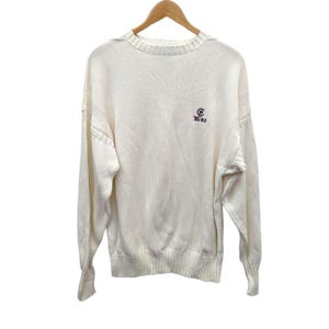 May include: Cream-colored knit sweater with long sleeves, a crew neck, and ribbed cuffs and hem. Features a small embroidered logo with the text "RL-83" in navy blue and red. The sweater is hanging on a wooden hanger.