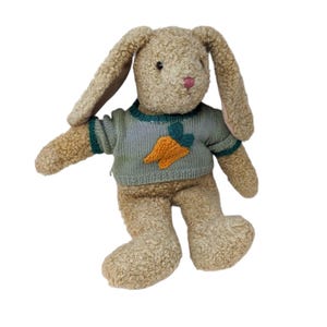 Puede incluir: Un peluche de conejito con orejas largas y una nariz rosa. El conejito lleva un suéter azul claro con una aplicación de zanahoria. El pelaje del conejito es de color marrón claro.