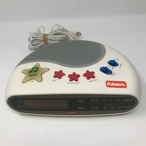 May include: White Playskool digital clock radio with a gray speaker and red, blue, and green buttons. The clock radio has a digital display with the words "FM" and "AM" and the frequencies "88 92 98 104 108 MHz" and "54 60 80 120 160 KHz".