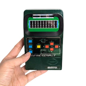 Puede incluir: Un juego de fútbol electrónico portátil vintage en una carcasa verde oscuro. El juego presenta una pequeña pantalla que muestra un campo de fútbol, con botones para puntuar, patear, pasar y correr. El texto "CLASSIC FOOTBALL 2" está impreso en la parte delantera, con el logotipo de Mattel.