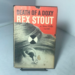 Pode incluir: Livro de bolso vintage intitulado "Death of a Doxy" de Rex Stout, um romance de Nero Wolfe. A capa apresenta um fundo vermelho com texto branco e um gráfico em preto e branco sobre uma imagem em tons de cinza de uma orelha e cabelo de mulher.